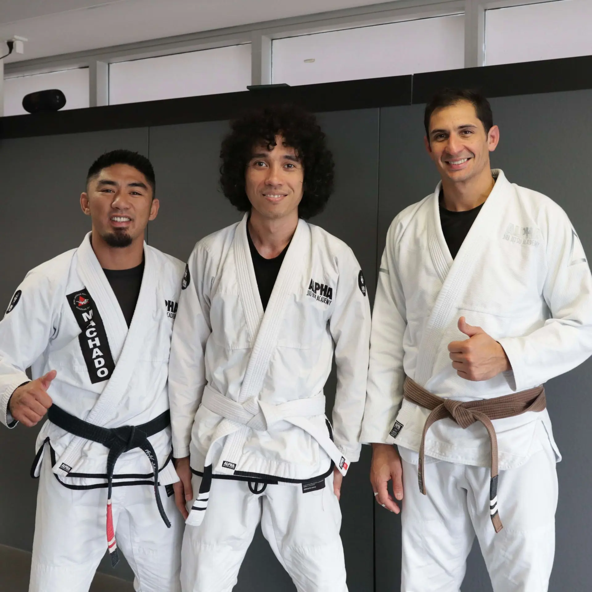 Three individuals, including two instructors and a student, stand together in their white gis at Alpha Jiu Jitsu Academy. The instructors, one wearing a black belt and the other with a brown belt, smile and give a thumbs-up, while the student proudly wears a white belt, signifying their progress in the martial art. This image captures the supportive atmosphere and camaraderie within the academy's training community.