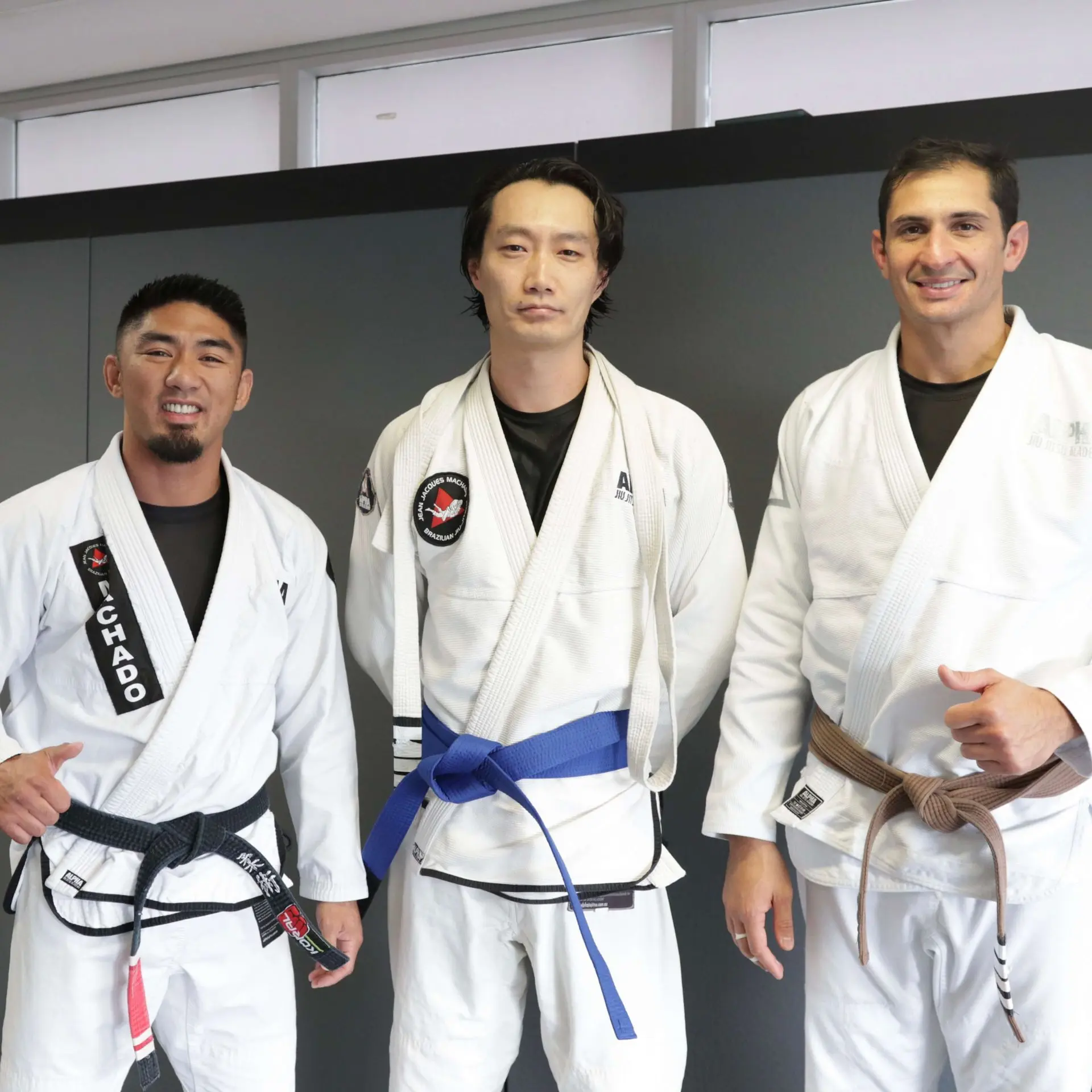 A student proudly displays their blue belt alongside two instructors at Alpha Jiu Jitsu Academy. The instructors, one wearing a black belt with "Machado" embroidery and the other a brown belt, stand on either side of the student, offering support and smiles. This image captures a moment of achievement and recognition in the Brazilian Jiu-Jitsu journey within the academy.