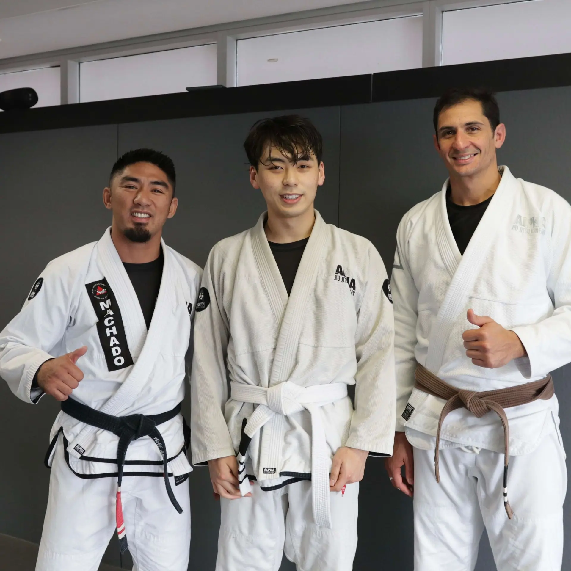 A white belt student stands between two experienced instructors at Alpha Jiu Jitsu Academy, smiling after a training session. The instructor on the left wears a black belt with "Machado" embroidered, while the instructor on the right wears a brown belt. This image captures a supportive moment of progress and camaraderie at the academy.