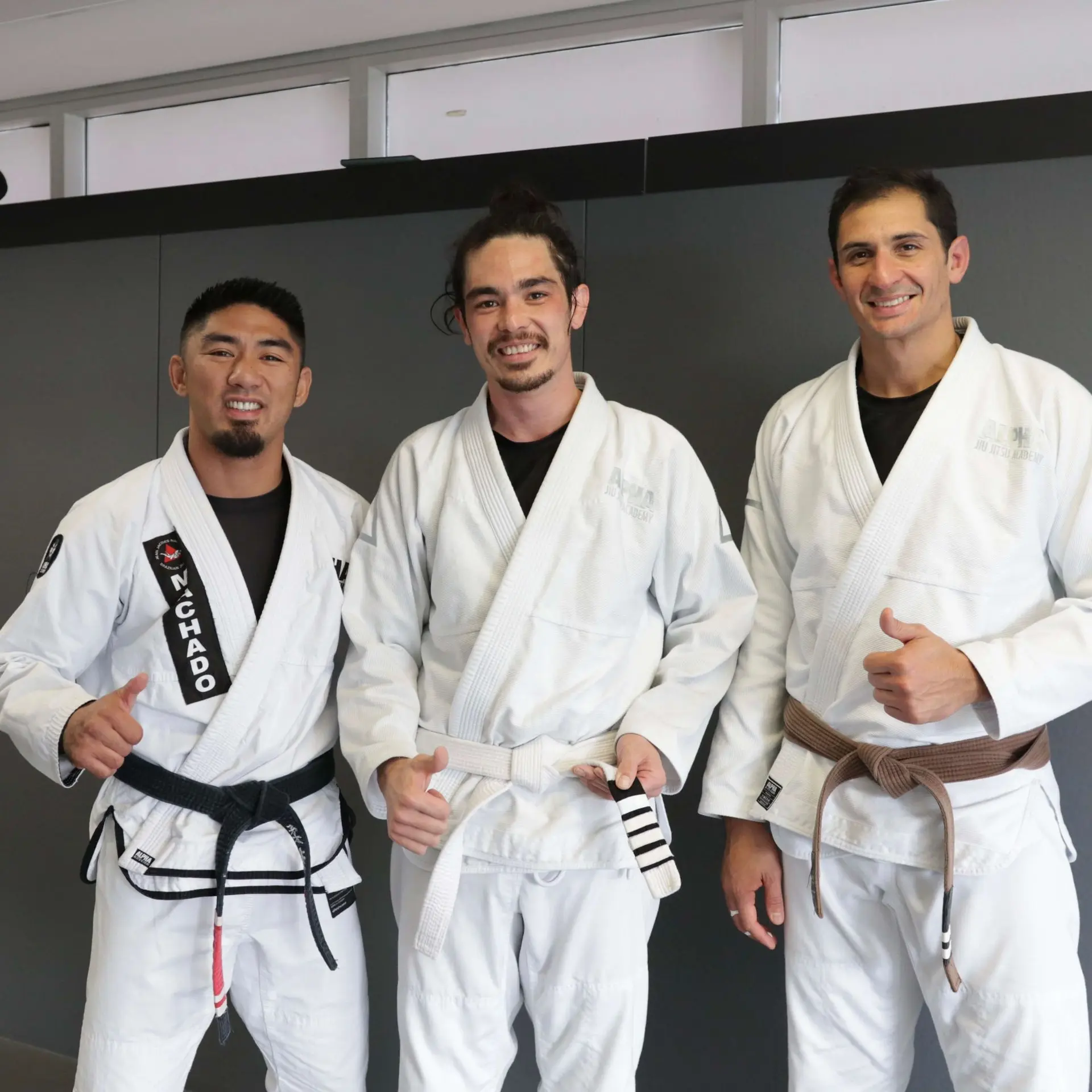 Three individuals pose together at Alpha Jiu Jitsu Academy, proudly wearing white gis. The central student holds their newly achieved belt stripe, flanked by two instructors. One instructor wears a black belt with "Machado" embroidered, and the other dons a brown belt, both offering smiles and supportive gestures. The image showcases the academy's emphasis on achievement and mentorship in the Brazilian Jiu-Jitsu journey.