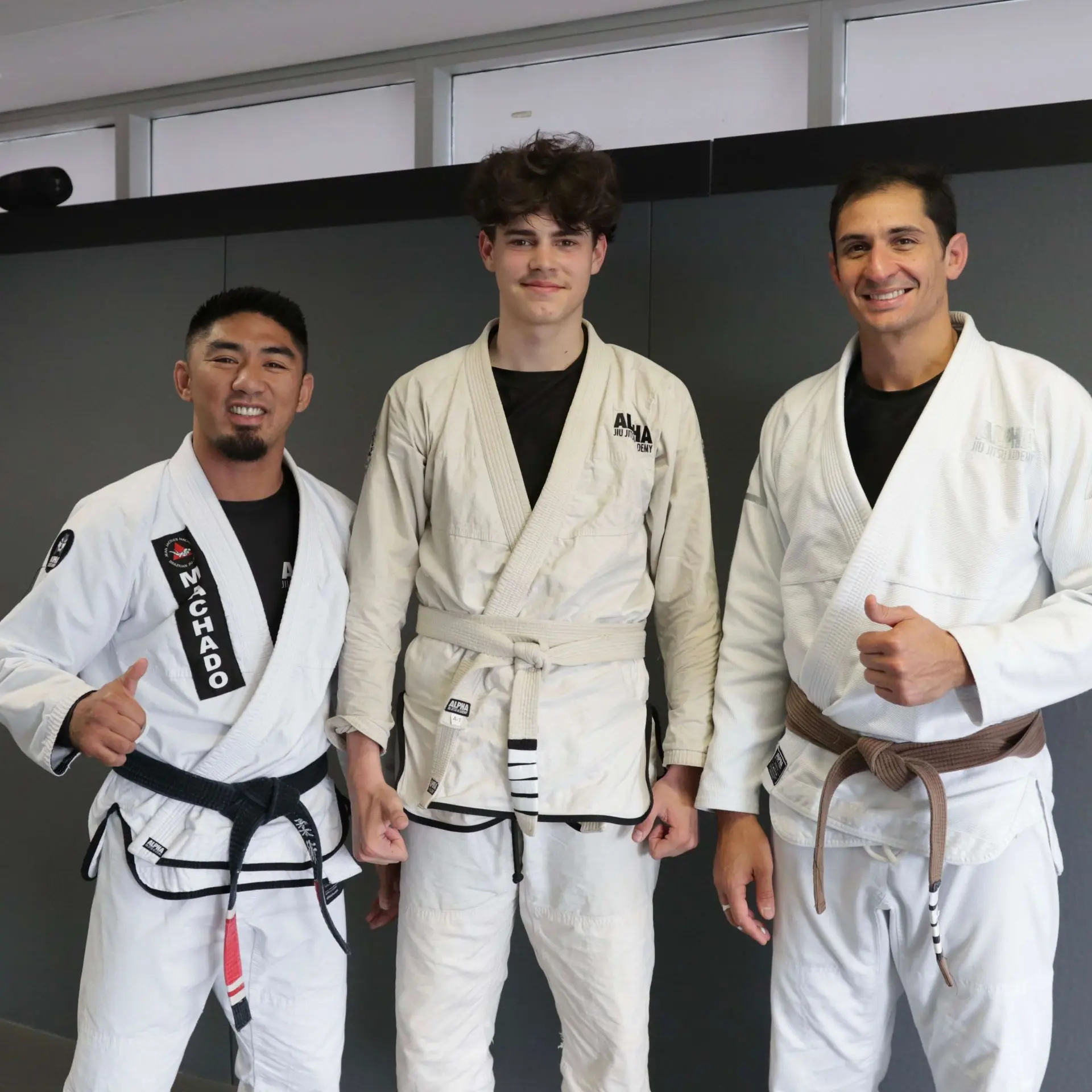 A young student wearing a white belt and a beige gi stands with two instructors at Alpha Jiu Jitsu Academy. The instructors, wearing black and brown belts, smile and give a thumbs-up, showcasing the supportive and encouraging environment at the academy.