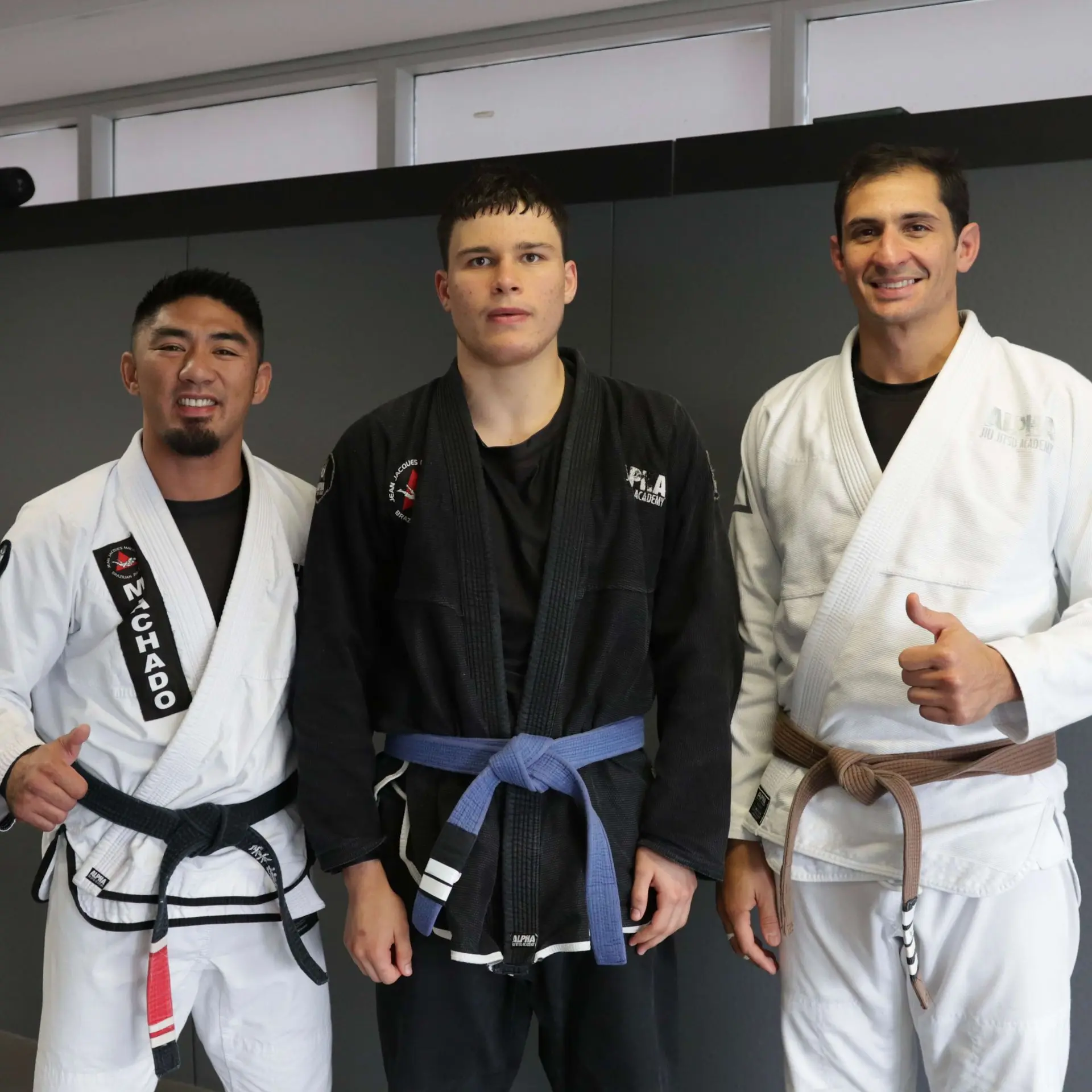 A blue belt student in a black gi stands with two instructors at Alpha Jiu Jitsu Academy. The instructors, wearing black and brown belts, pose beside the student, with one giving a thumbs-up, reflecting the supportive and skill-focused training environment of the academy.