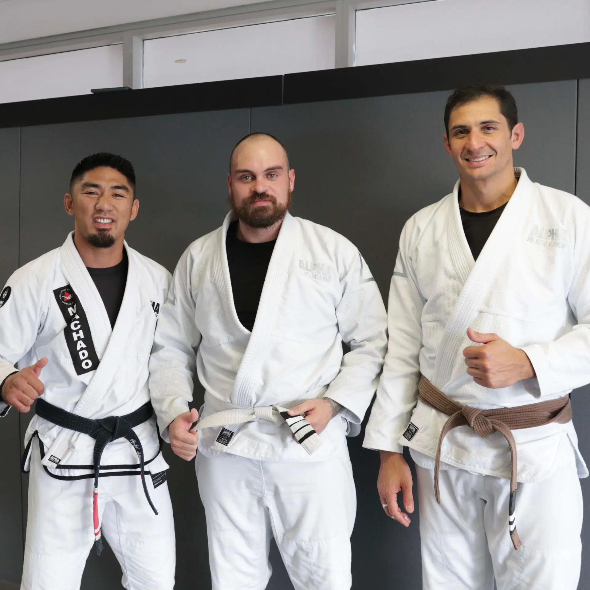 A student wearing a white belt stands between two instructors at Alpha Jiu Jitsu Academy, all dressed in white gis, with the instructors wearing black and brown belts, posing together with smiles and a thumbs-up, symbolizing mentorship and training achievements at the academy.