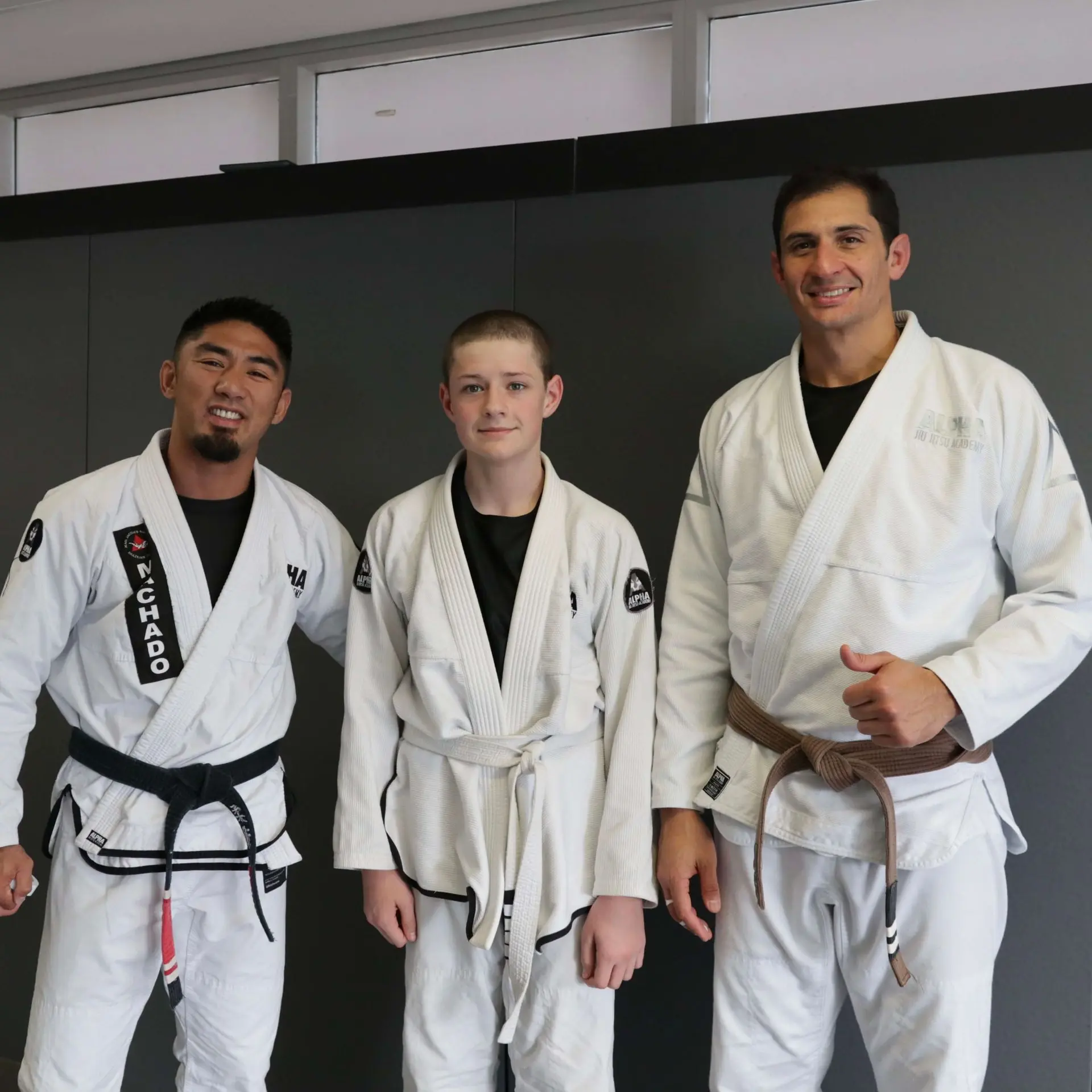 A white belt student stands between two instructors from Alpha Jiu Jitsu Academy, both wearing white gis with brown and black belts, posing together with smiles and a thumbs-up, representing mentorship and training progress at the academy.