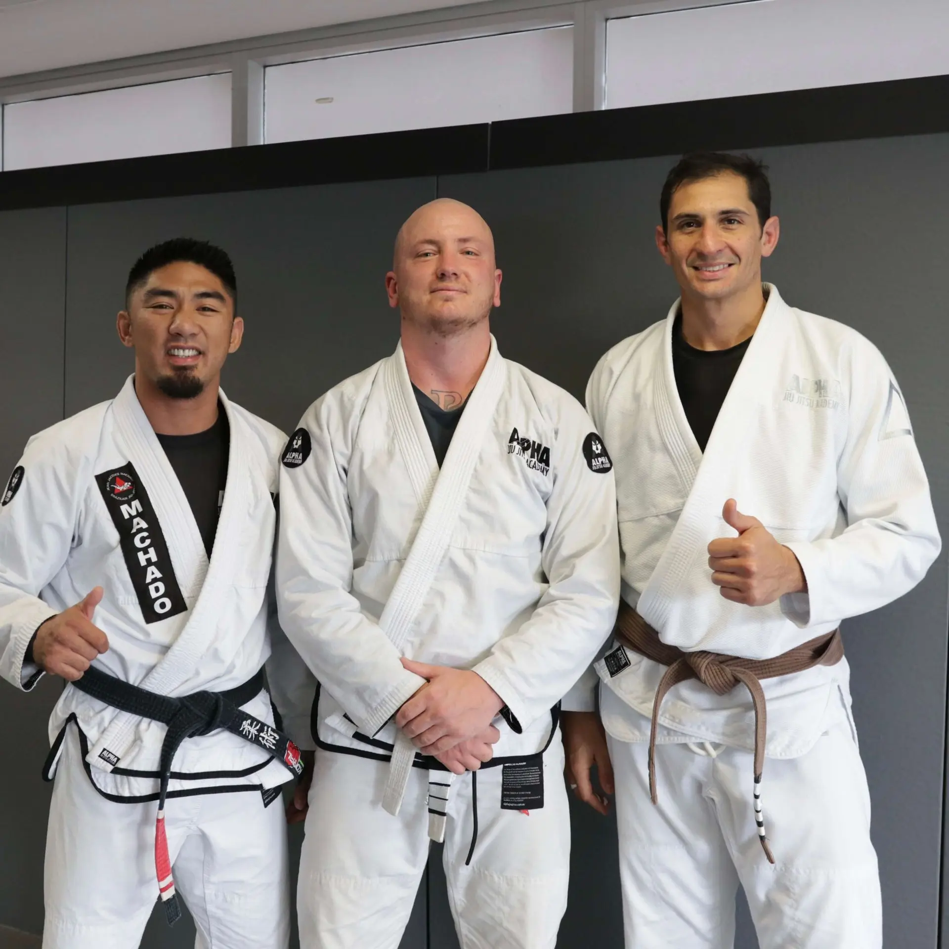 A student proudly poses after receiving his white belt at Alpha Jiu Jitsu Academy, standing with two instructors. The instructor on the left is wearing a black belt, and the instructor on the right is wearing a brown belt, both giving thumbs-up in celebration. The student is wearing a freshly earned white gi, symbolizing the start of their Jiu Jitsu journey. The academy's branding is visible on the gis. The image is captured with a neutral grey backdrop, creating a clean and professional look.