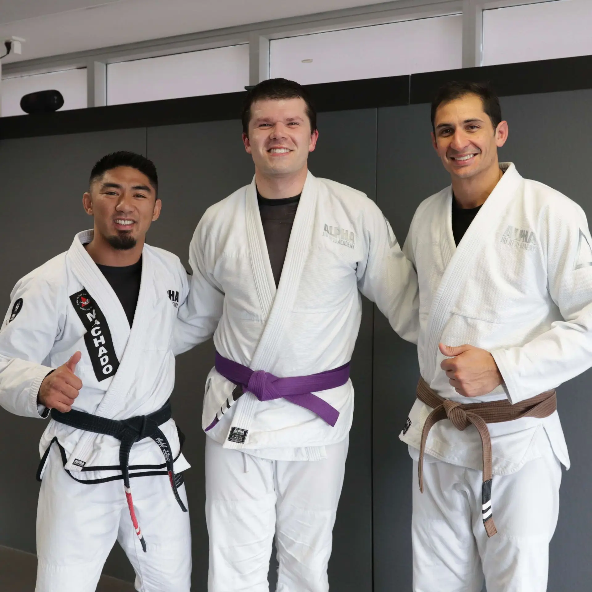 A student proudly stands with two instructors after receiving his purple belt at Alpha Jiu Jitsu Academy. The instructor on the left is wearing a black belt, and the instructor on the right is wearing a brown belt. The student, now a purple belt, smiles broadly as he celebrates this milestone in his Jiu Jitsu journey. Both instructors are giving thumbs-up in recognition of the achievement, and the academy's branding is visible on their gis. The background is a neutral grey, creating a clean and professional atmosphere.