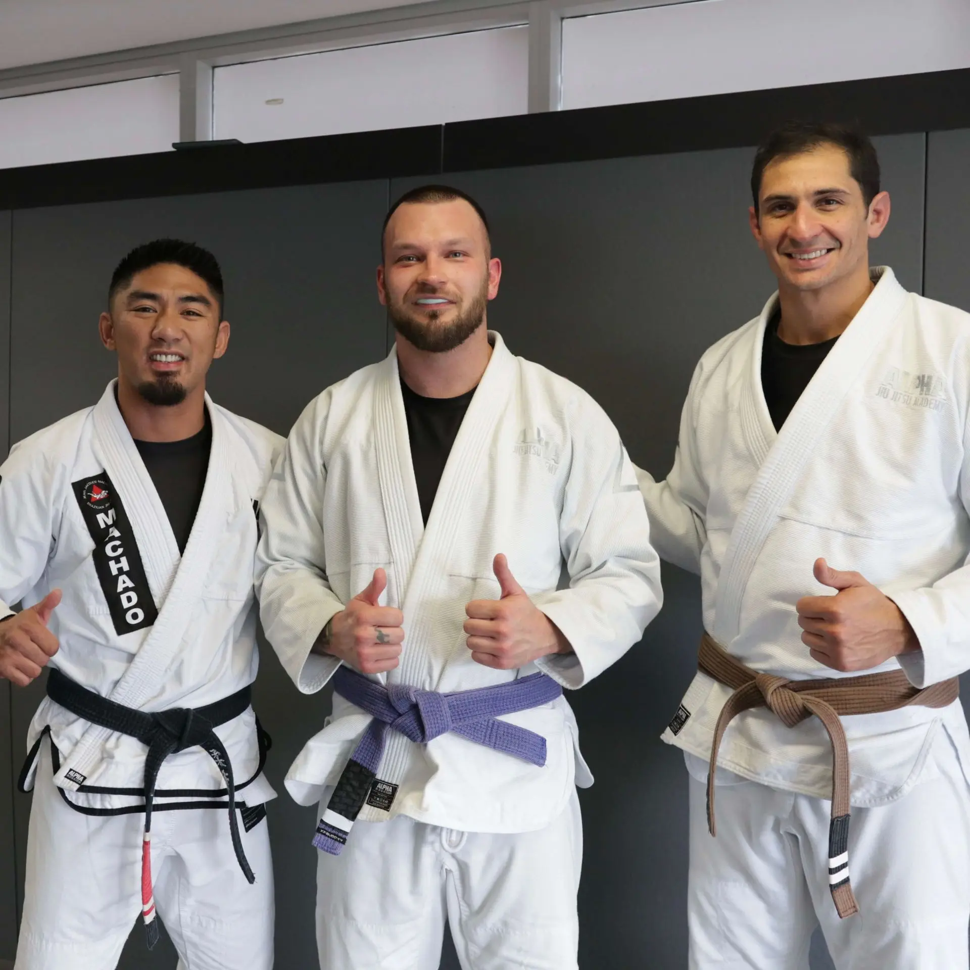 A student proudly stands in the middle wearing a newly awarded purple belt alongside two instructors from Alpha Jiu Jitsu Academy, all giving a thumbs-up gesture. All three are dressed in white gis with black belts and patches, reflecting a supportive moment during a belt promotion ceremony at the academy. The scene captures smiles and camaraderie in front of a grey training backdrop.