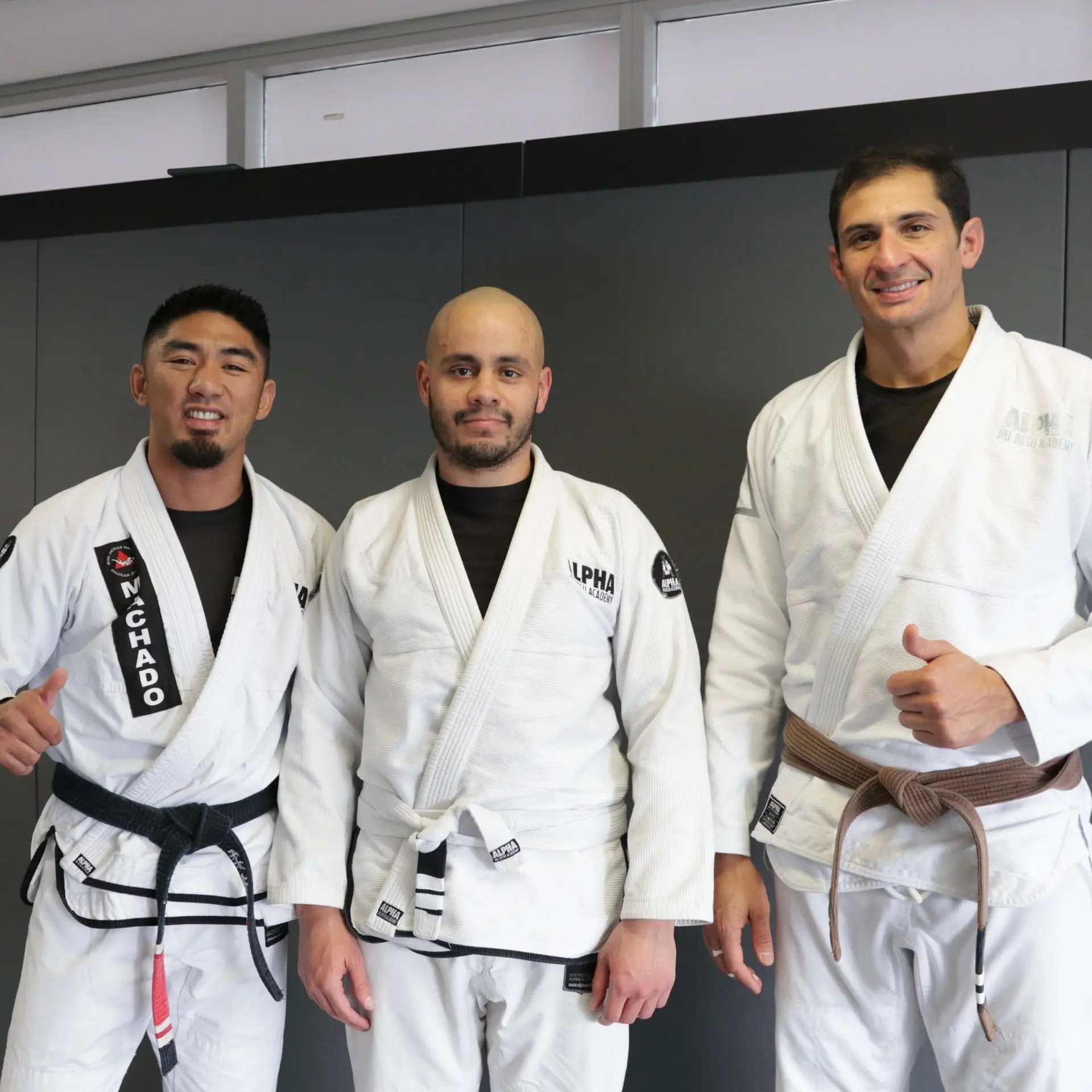 A proud student stands between two instructors at Alpha Jiu Jitsu Academy, celebrating his purple belt promotion. The student, wearing a white gi and his new purple belt, smiles alongside the instructors, one wearing a black belt and the other a brown belt. The photo captures a joyful moment with all three giving thumbs-up in front of a neutral grey backdrop. The academy's logo is visible on their gis, highlighting the spirit of achievement and camaraderie.