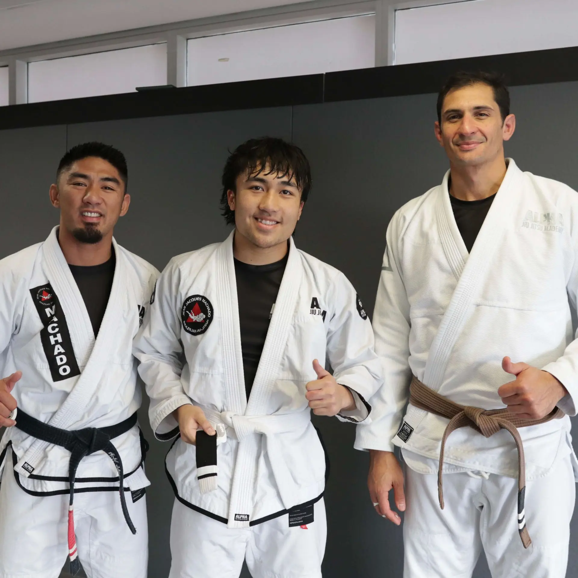 A photo showing three individuals in a martial arts studio. On the left, a male instructor with a black belt in a white gi gives a thumbs up. In the center, a male student in a white gi with a white belt smiles while holding his belt. On the right, another instructor with a brown belt in a white gi gives a thumbs up. The background shows a gray wall in the academy.