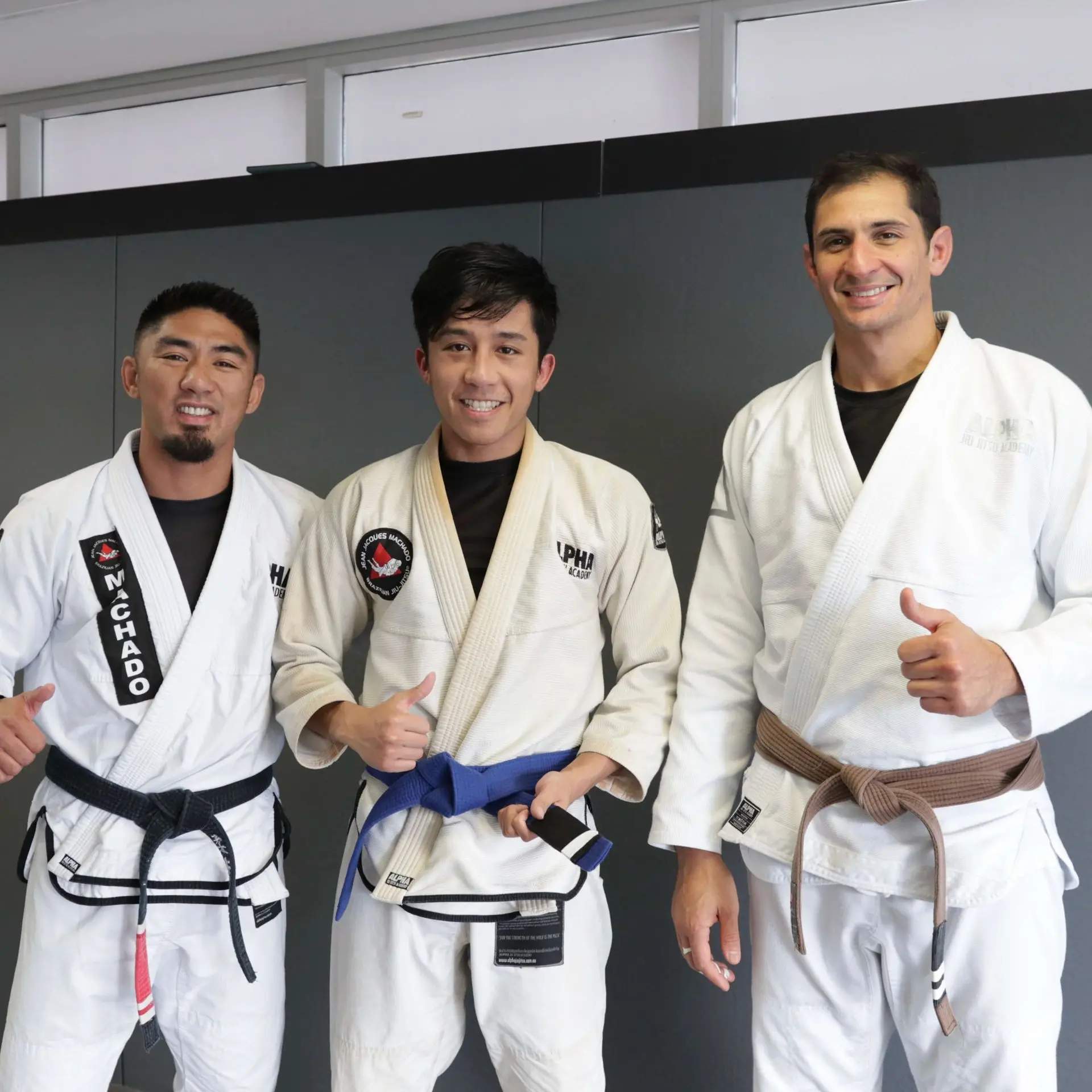 A photo showing three individuals in a martial arts academy. On the left, a male instructor in a black belt with a white gi gives a thumbs up. In the center, a male student in a white gi with a blue belt smiles while holding his belt. On the right, another instructor with a brown belt in a white gi also gives a thumbs up. The background features a gray wall.