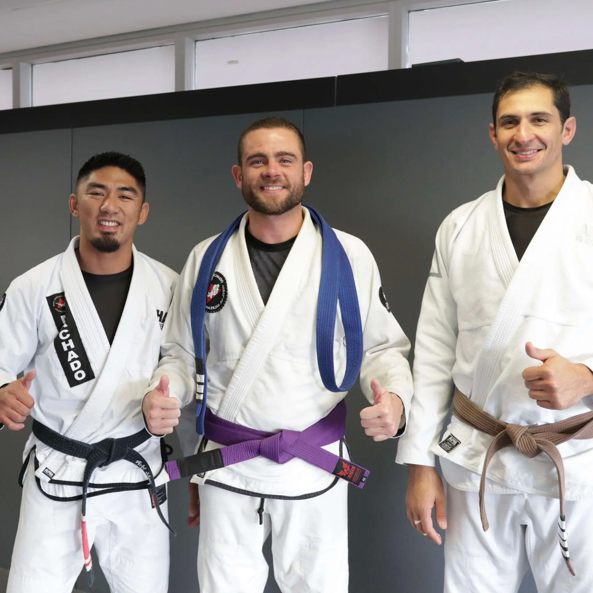 A purple belt student in a white gi stands between two instructors at Alpha Jiu Jitsu Academy. Both instructors are wearing black and brown belts, with the group giving thumbs-up gestures, showcasing camaraderie and the positive training atmosphere within the academy.