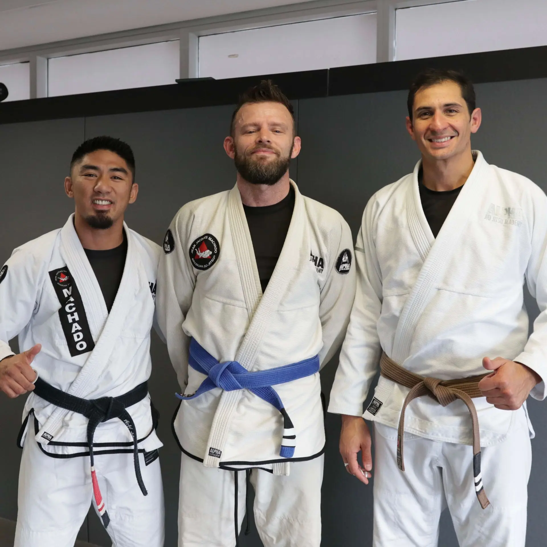 A photo of three individuals in a Jiu-Jitsu academy. On the left, a male instructor in a black belt with a white gi gives a thumbs up. In the center, a male student wearing a blue belt and white gi stands with a serious expression. On the right, another instructor in a brown belt with a white gi smiles while giving a thumbs up. The background features a simple gray wall.