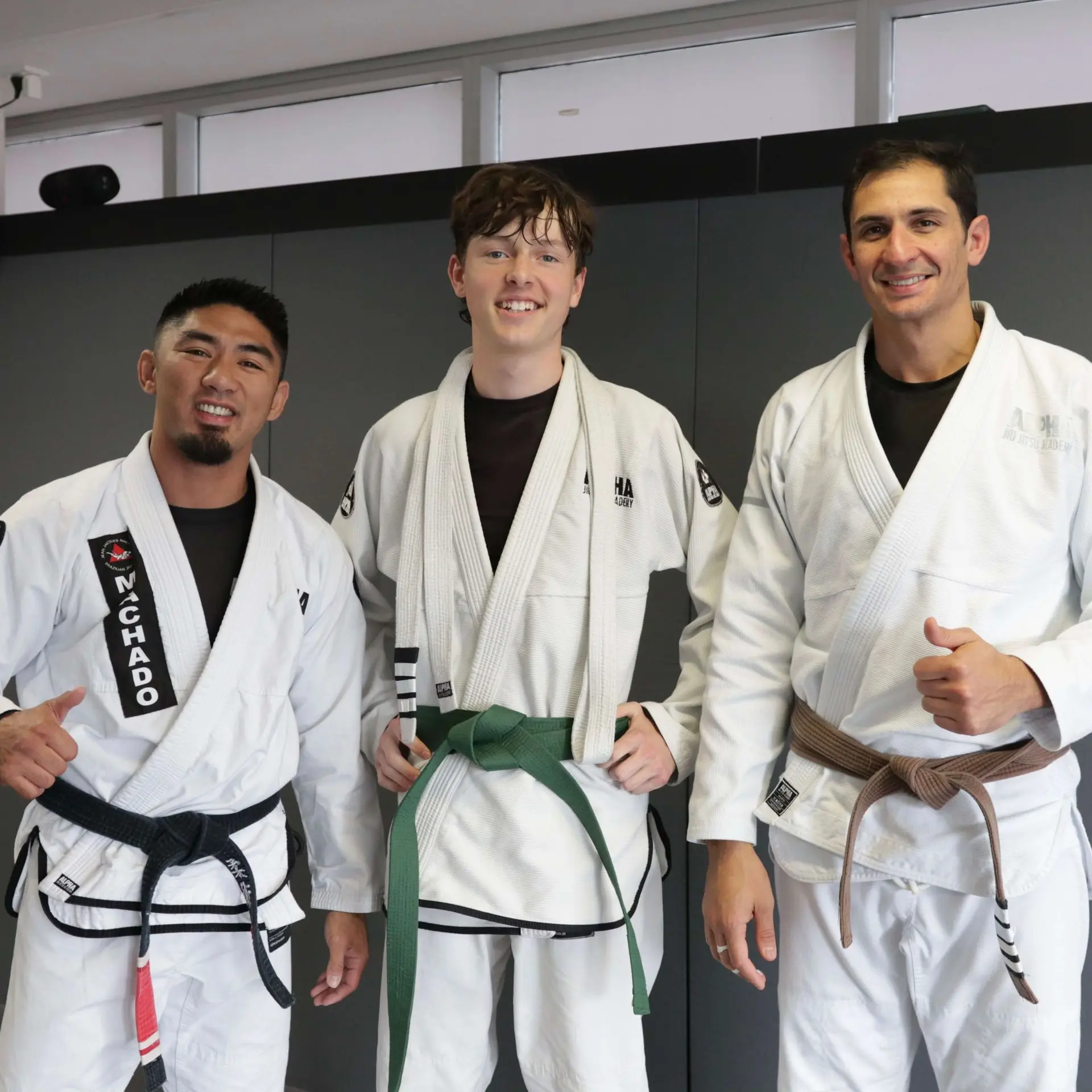 A student proudly wearing a green belt stands with two instructors at Alpha Jiu Jitsu Academy. The instructor on the left, wearing a black belt embroidered with "Machado," and the instructor on the right, wearing a brown belt, pose with the student for a photo. This image highlights progress and mentorship within the jiu-jitsu training environment.