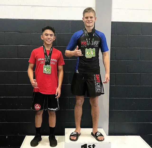 Alpha Jiu Jitsu Academy competitor standing on the first-place podium next to another medal-winning athlete after a grappling competition, both showing thumbs-up.