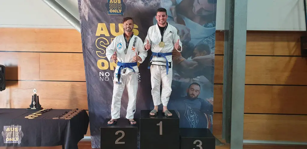 Two competitors standing on the 1st and 2nd place podiums at the AUS Sub Only competition, both wearing blue belts and gi uniforms, smiling and holding their medals. The backdrop features the event branding and a large promotional banner.