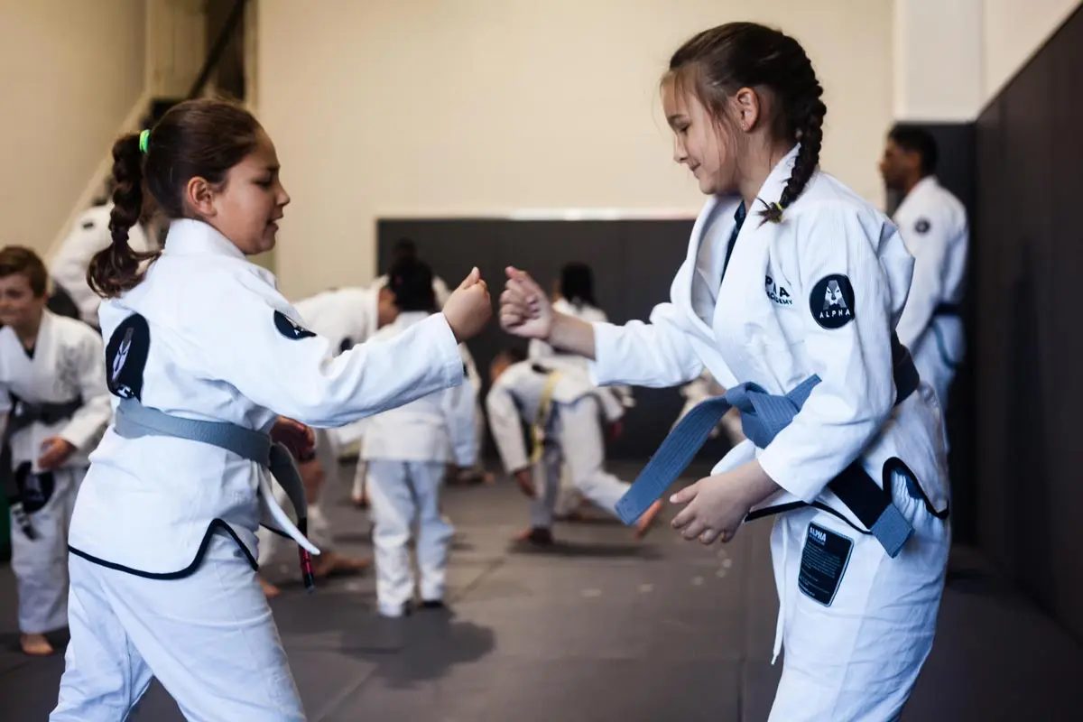 Two young students in white gis and gray belts from Alpha Jiu Jitsu Academy engage in a friendly training exercise on the mats, surrounded by other students practicing in the background. Both students are smiling and preparing to grapple, showcasing a positive and engaging atmosphere during their training session.