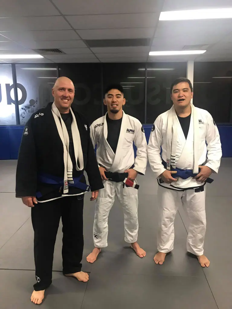 Three jiu-jitsu practitioners, including an instructor from Alpha Jiu Jitsu Academy, standing side by side on the academy's mats, all wearing blue belts and gis, with two in white gis and one in a black gi, smiling at the camera during a training session.