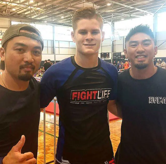 Two coaches from Alpha Jiu Jitsu Academy pose alongside a competitor wearing a "Fight Life" rash guard at a jiu-jitsu competition venue. All three are smiling, capturing a moment of camaraderie and support during the event, with a busy indoor sports setting in the background.Two coaches from Alpha Jiu Jitsu Academy pose alongside a competitor wearing a "Fight Life" rash guard at a jiu-jitsu competition venue. All three are smiling, capturing a moment of camaraderie and support during the event, with a busy indoor sports setting in the background.