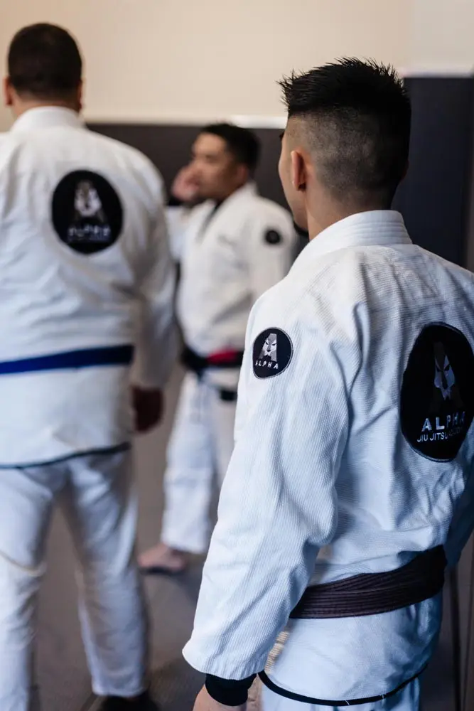 Xavier Sales, Alpha Jiu Jitsu instructor, wearing a gi with the Alpha Jiu Jitsu Academy logo visible on the back, overseeing a class in session, with other students blurred in the background.