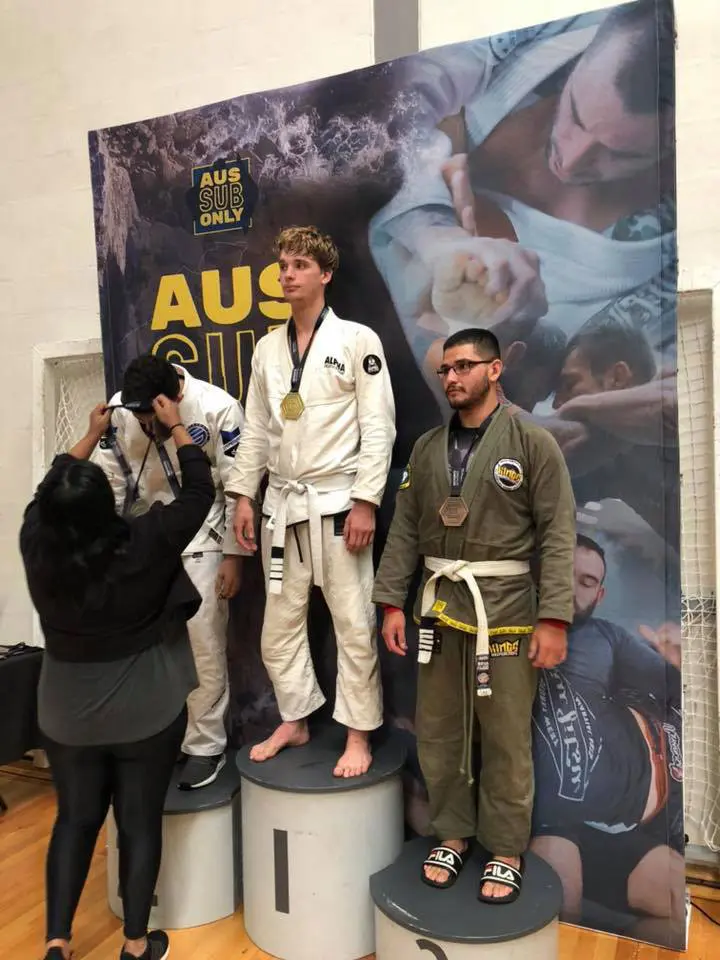 Competitors standing on the podium at the AUS SUB Only Championship, with the first-place winner from Alpha Jiu Jitsu Academy wearing a white gi and gold medal. Other medalists in second and third places stand on the podium in their respective positions, while a presenter adjusts a medal on one of the participants. The backdrop features the event branding.