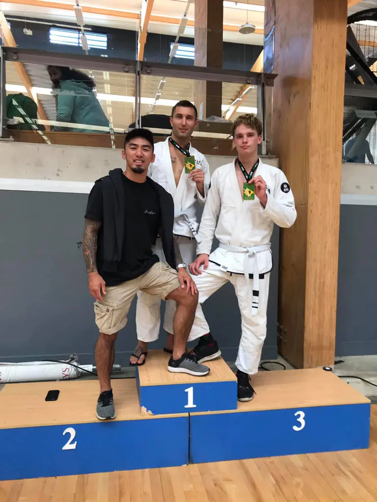 Two Alpha Jiu Jitsu Academy students standing on a podium in first and third place, proudly displaying medals, accompanied by their coach Xavier Sales wearing casual attire at a competition venue.