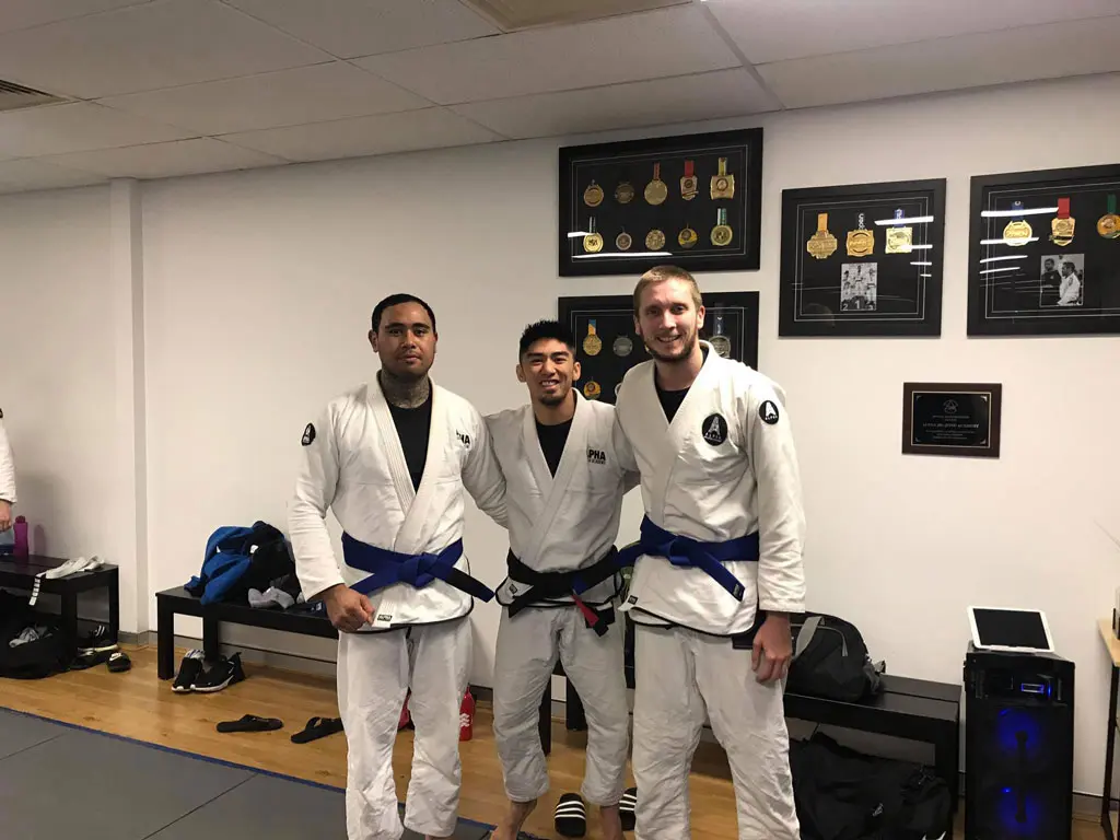 Three martial artists from Alpha Jiu Jitsu Academy wearing white gis and blue belts posing together with smiles in front of a wall displaying framed medals and awards, showcasing their achievements and camaraderie.