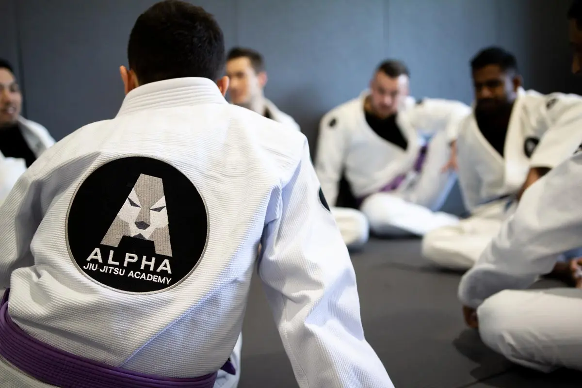 A group of jiu-jitsu practitioners in gi uniforms seated in a circle, with the focus on a person wearing a gi displaying the Alpha Jiu Jitsu Academy logo prominently on the back. The atmosphere is casual and conversational, with students discussing or preparing for training.