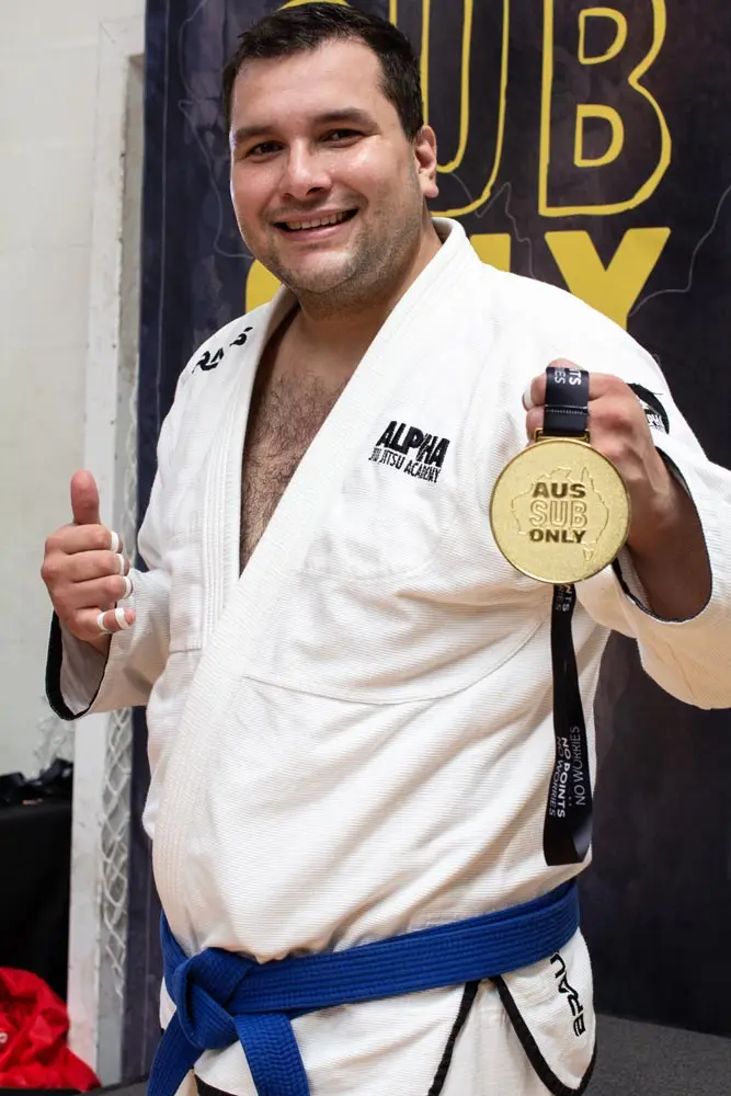 Alpha Jiu Jitsu Academy competitor smiling and holding a gold medal at a Brazilian Jiu Jitsu competition, highlighting achievement and dedication.