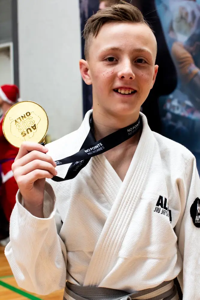 Youth student from Alpha Jiu Jitsu Academy proudly holding a gold medal after winning a BJJ competition, wearing a white gi and gray belt.