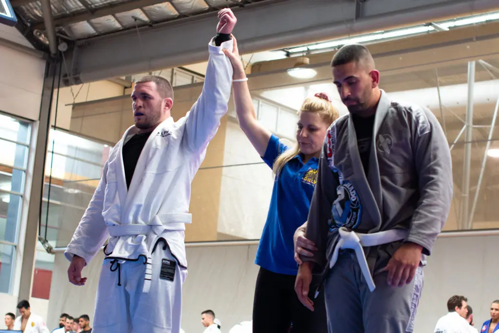 An Alpha Jiu Jitsu Academy competitor in a white gi has their hand raised by a referee in a blue shirt, signifying victory, while their opponent in a gray gi stands to the right with their head down. The scene takes place inside a brightly lit indoor gymnasium with other onlookers in the background.