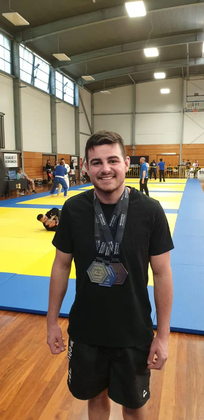 A smiling competitor from Alpha Jiu Jitsu Academy wearing a black shirt and shorts, standing on a gymnasium floor with two medals around his neck. The background shows a Brazilian Jiu-Jitsu competition taking place, with mats and other participants visible.