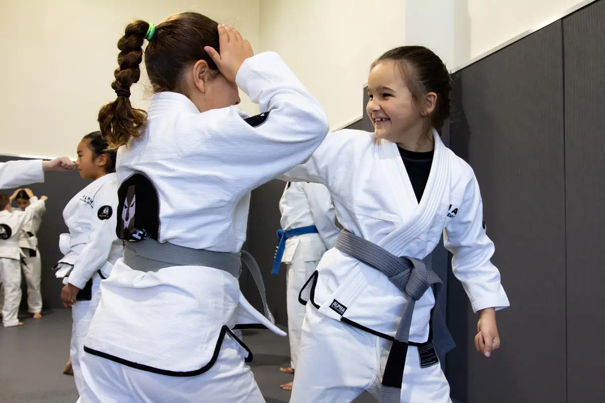 Two young girls practicing jiu-jitsu techniques at Alpha Jiu Jitsu Academy, wearing white gis with gray belts, engaging in playful training with smiles and focus. Other students in white gis are seen practicing in the background on the academy's training mats.