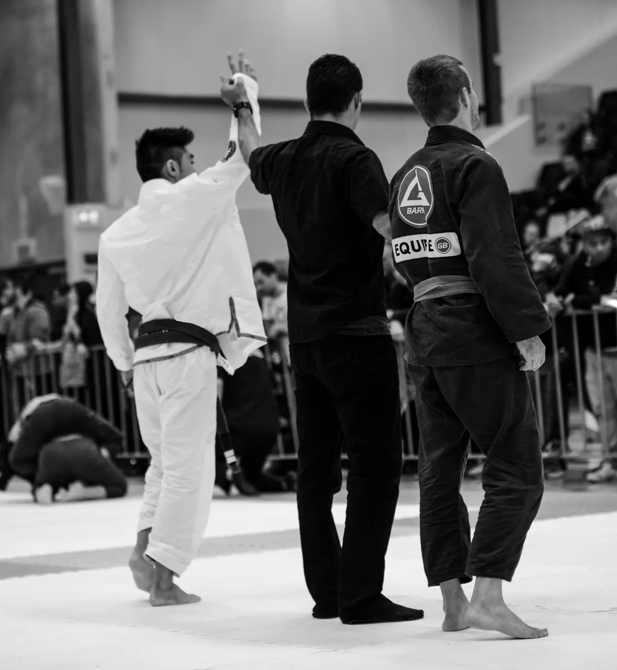 Black and white image capturing a victorious Alpha Jiu Jitsu Academy competitor in a white gi as their hand is raised by a referee, while their opponent in a dark gi stands on the right. The scene unfolds in a crowded gymnasium with spectators watching from behind barriers.