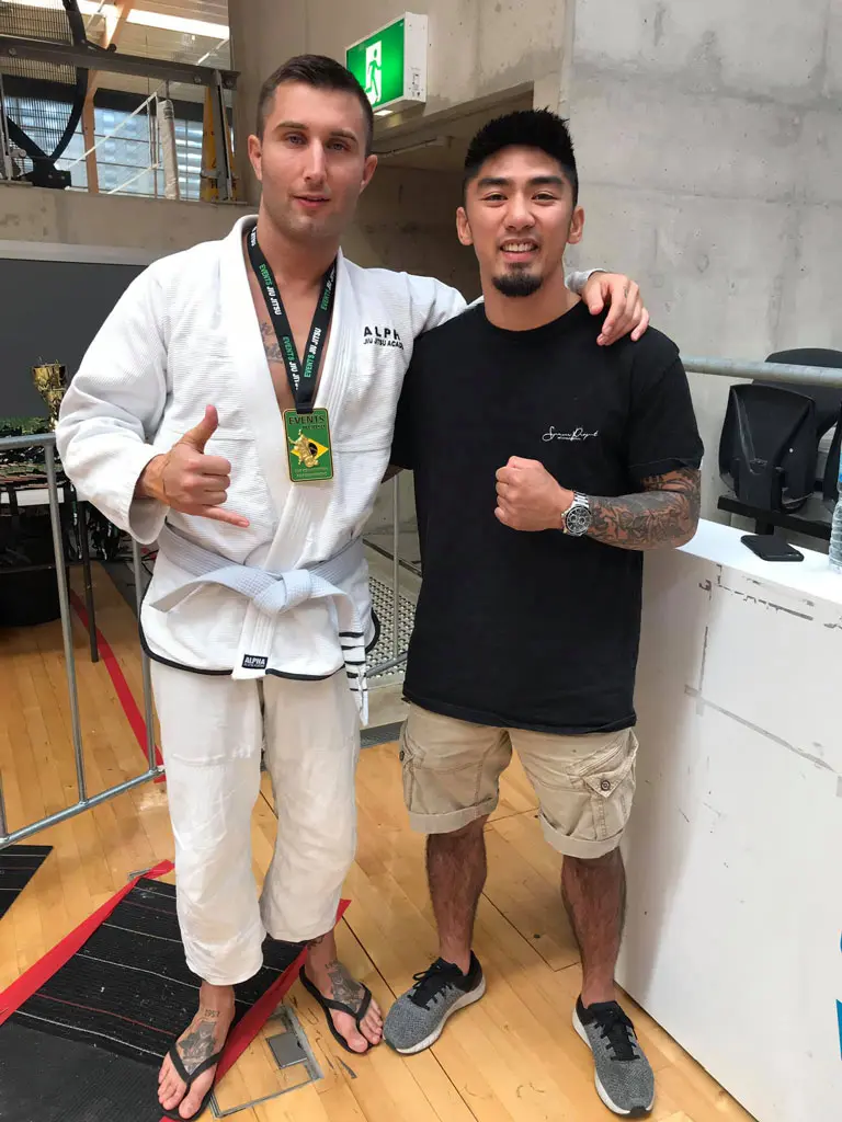 A jiu-jitsu competitor from Alpha Jiu Jitsu Academy wearing a white gi with a green medal around his neck stands next to his coach, who is wearing a black T-shirt and khaki shorts, posing for a photo at a competition venue. Both are smiling and showing a gesture of camaraderie.