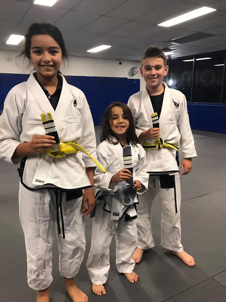 Three young students at Alpha Jiu Jitsu Academy celebrating their belt promotion, proudly holding their new belts while smiling on the training mats.