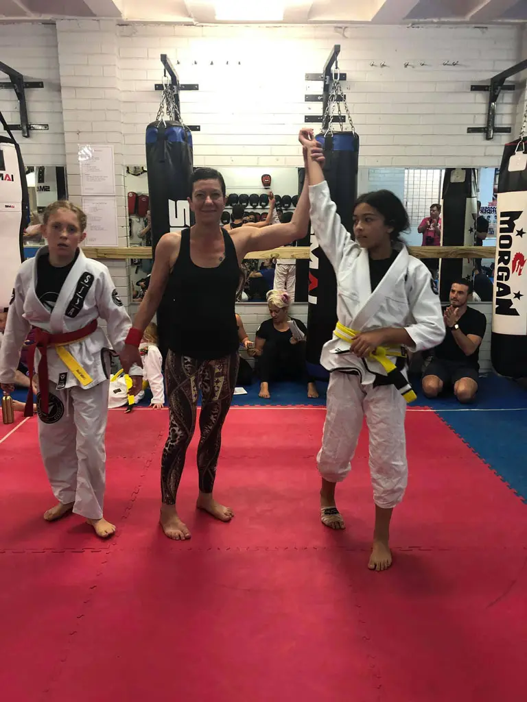 A young competitor from Alpha Jiu Jitsu Academy wearing a white gi and yellow belt has their hand raised in victory by a referee. The scene is set in a gym with punching bags and other spectators in the background, while another young competitor stands to the left, looking towards the winner.