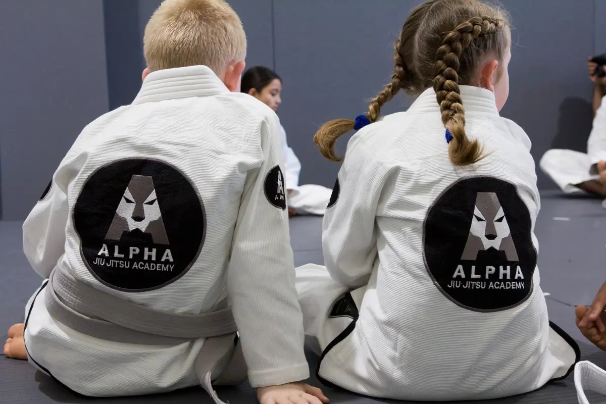 Young boy and girl sitting in white gis with gray belts, displaying the Alpha Jiu Jitsu Academy logo on their backs, attentively participating in training at a martial arts session.