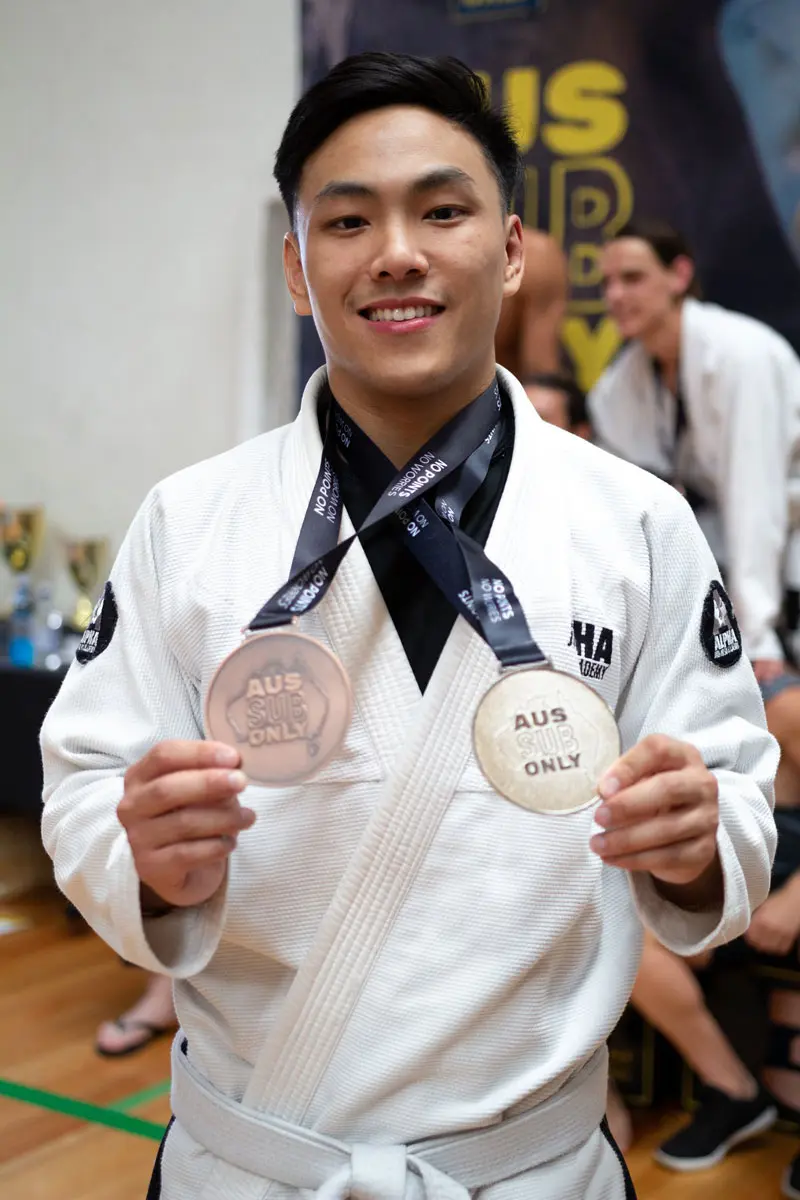 Smiling Alpha Jiu Jitsu Academy student proudly displaying two AUS SUB ONLY medals while wearing a white gi with Alpha Jiu Jitsu branding.