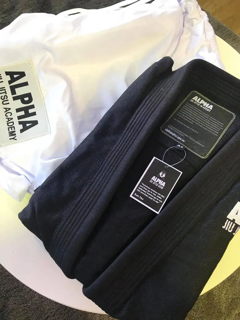 Alpha Jiu Jitsu Academy branded black and white gi uniforms displayed with a tag and academy details.