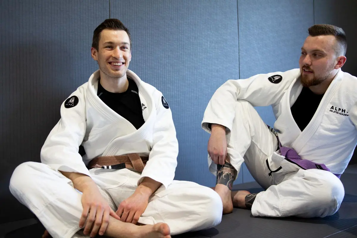 Two Alpha Jiu Jitsu Academy students, one wearing a brown belt and the other a purple belt, seated and smiling during a break at the academy.