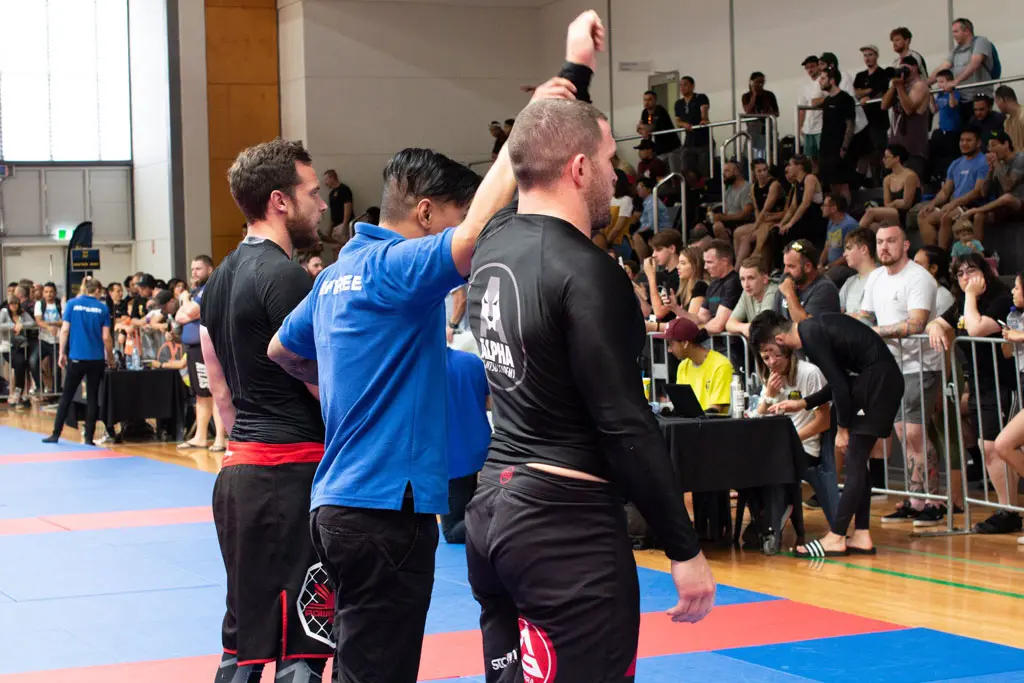 Alpha Jiu Jitsu Academy competitor being declared winner during a grappling competition, with referee raising the athlete's arm in victory.