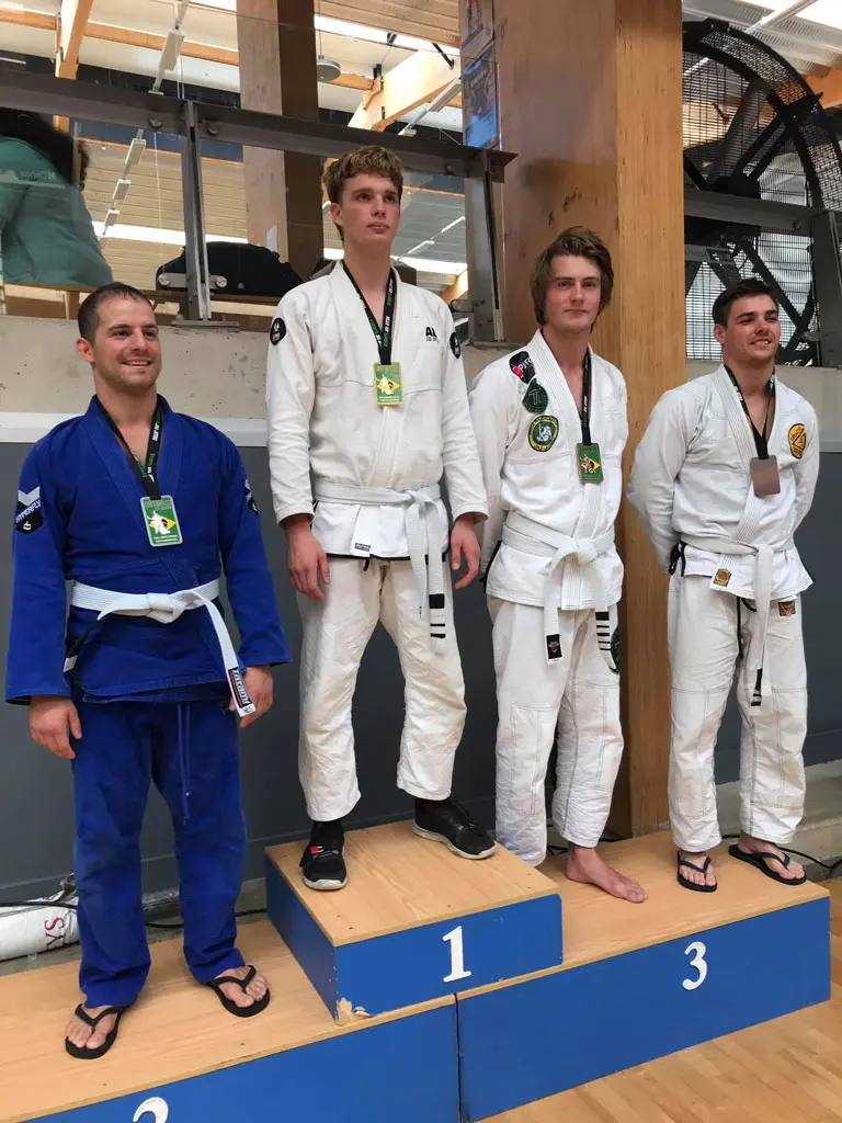 An Alpha Jiu Jitsu Academy athlete stands on the top spot of the podium wearing a white gi and a green medal at a jiu-jitsu competition. He is flanked by three other competitors, one in a blue gi and two in white gis, each displaying medals, standing on the second and third-place podiums. The setting is an indoor competition venue.