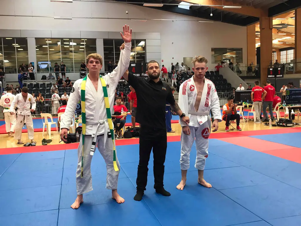 Alpha Jiu Jitsu competitor has his hand raised in victory by a referee after a match, with both competitors wearing gi uniforms on a blue and red mat in a competition venue.