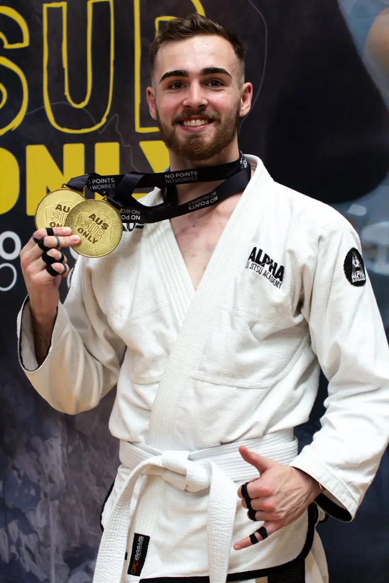 Alpha Jiu Jitsu Academy white belt competitor proudly displaying two gold medals from the AUS Sub Only competition, smiling in front of event backdrop.