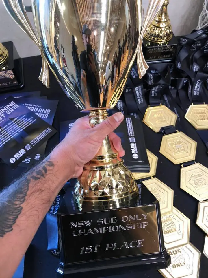 Hand holding a large golden trophy awarded for 1st place at the NSW Sub Only Championship, surrounded by medals and certificates.