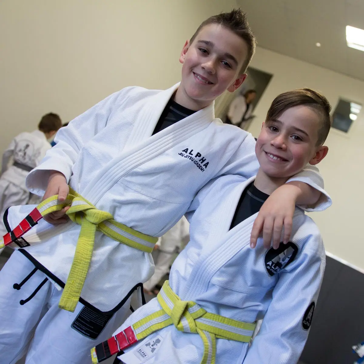 Two young students at Alpha Jiu Jitsu Academy wearing white gi uniforms and yellow belts smile while posing together, one arm around each other’s shoulder, inside the academy training room. The background shows other students engaged in activities.