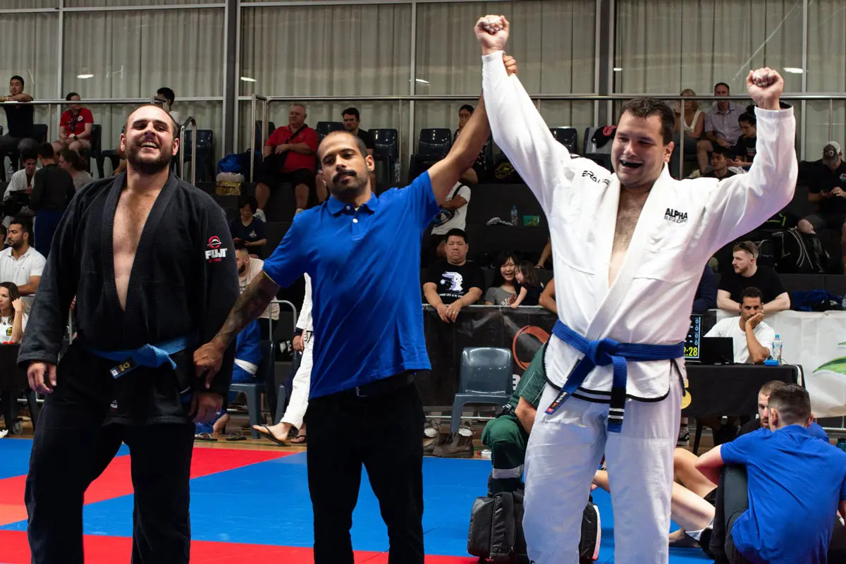 Alpha Jiu Jitsu competitor has his hand raised in victory by a referee after a match, with both competitors wearing gi uniforms on a blue and red mat in a competition venue.