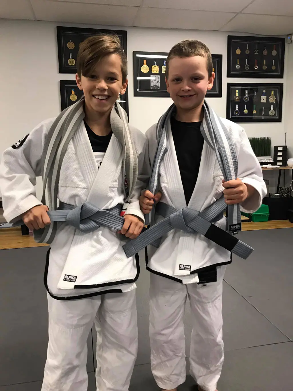 Two young students from Alpha Jiu Jitsu Academy stand proudly with their new belts draped over their shoulders after a promotion ceremony. Both are wearing white gi uniforms and smiling, with a wall of framed medals and awards displayed behind them.