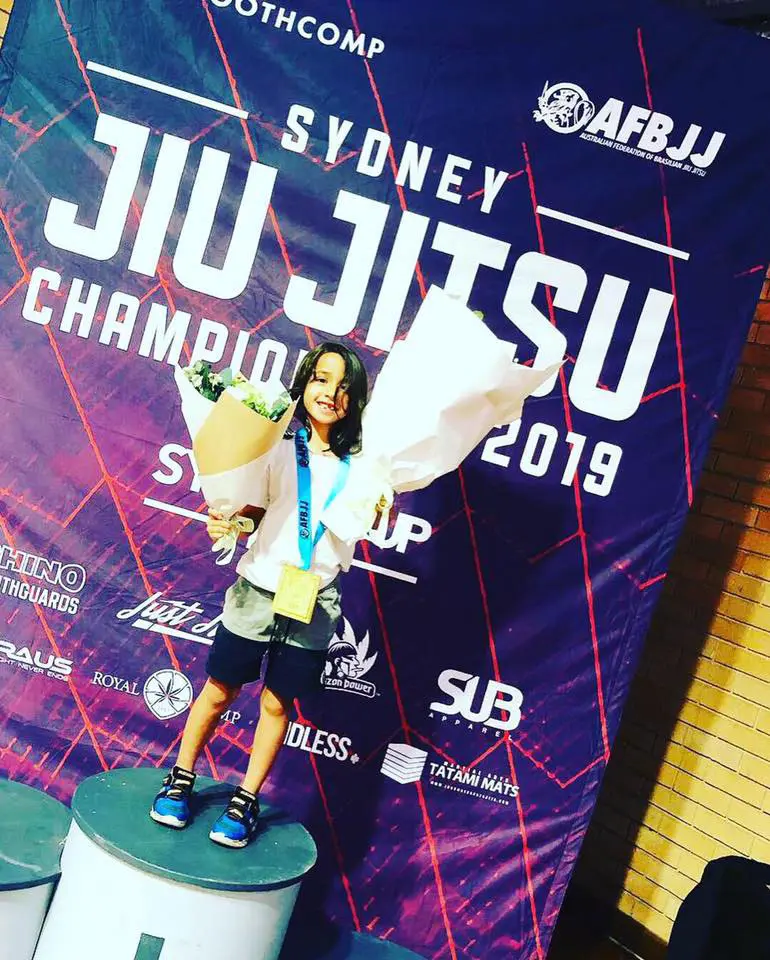 A young Alpha Jiu Jitsu Academy athlete stands on a podium holding a bouquet of flowers and wearing a gold medal around their neck at the Sydney Jiu Jitsu Championship 2019. The backdrop displays the event branding with prominent text reading "Sydney Jiu Jitsu Championship 2019." The athlete is smiling and celebrating the win.